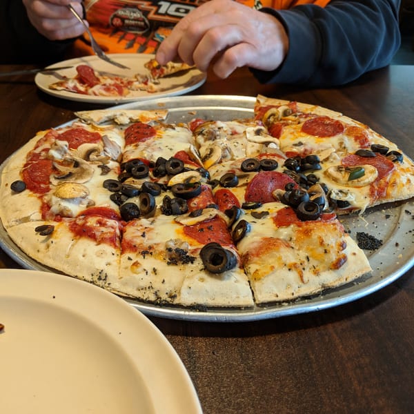 A person in an orange shirt eating a slice of pepperoni, mushroom, and olive pizza.