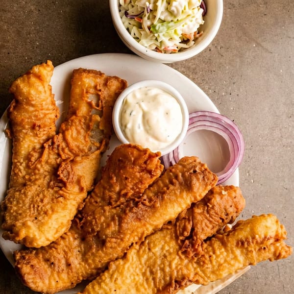 Golden-brown fried fish fillets on a white plate with tartar sauce, red onion, and coleslaw.