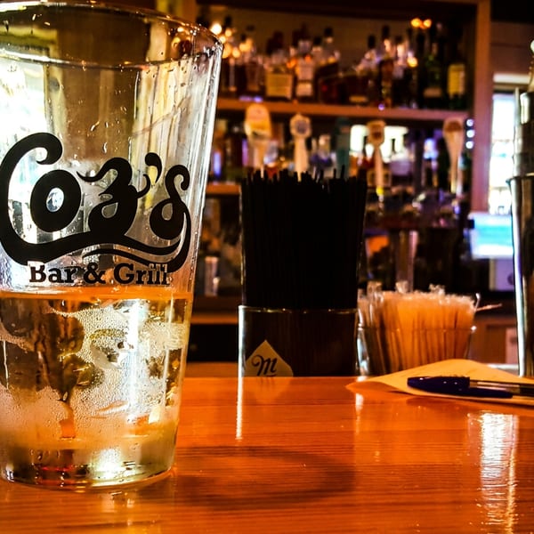 A Coz's Bar & Grill pint glass with a drink sits on a wooden bar counter.