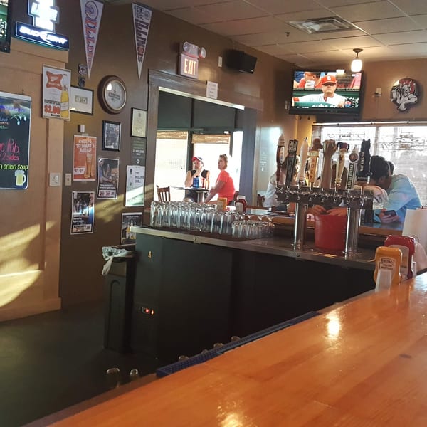 A casual bar interior with a wooden counter, beer taps, TVs, and patrons seated at tables.
