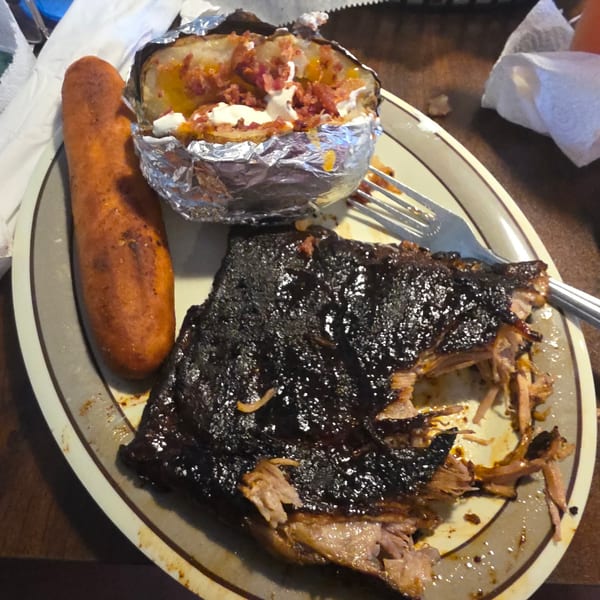 A plate of BBQ ribs, a loaded baked potato in foil, and a corn dog.