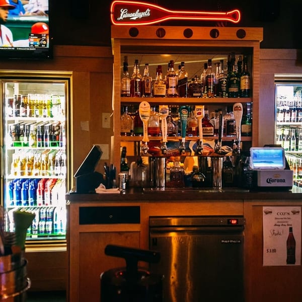 A cozy bar interior with a well-stocked liquor shelf, beer taps, and two TVs showing a baseball game.